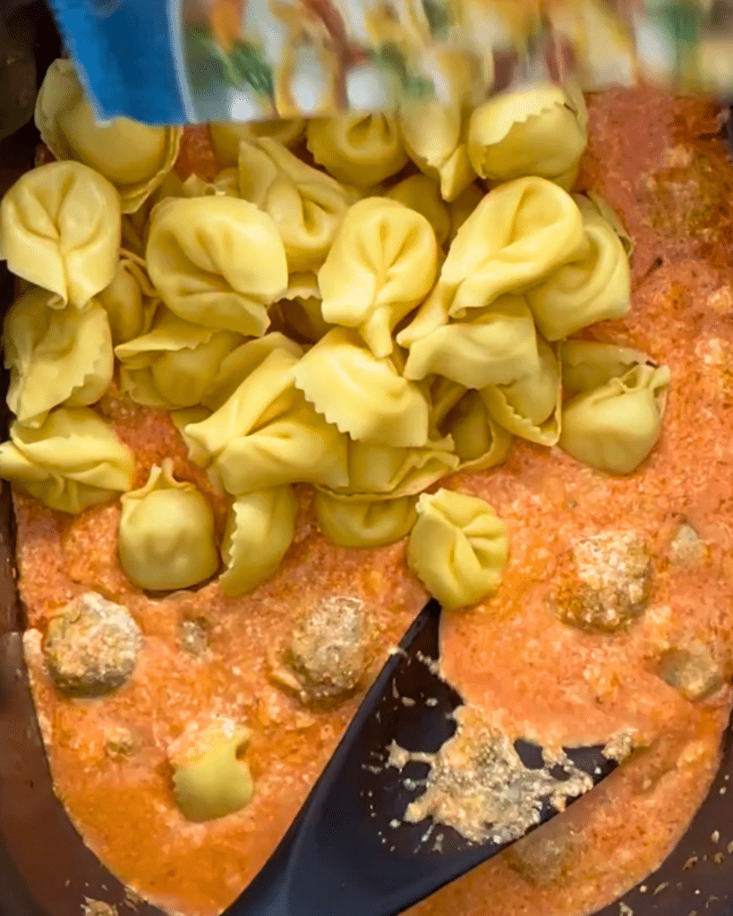 A black spoon stirs cheese tortellini into creamy buffalo sauce with meatballs in a pot, preparing a pasta dinner.