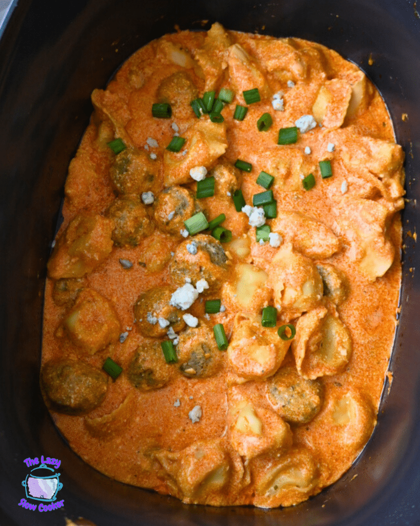 Ravioli and meatballs in a slow cooker are coated in creamy orange buffalo sauce, topped with green onions and cheese. "The Lazy Slow Cooker" logo appears.