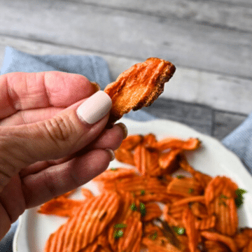 A hand with pale nail polish lifts a crinkle-cut carrot chip over a herb-garnished bowl of chips on a wooden table.