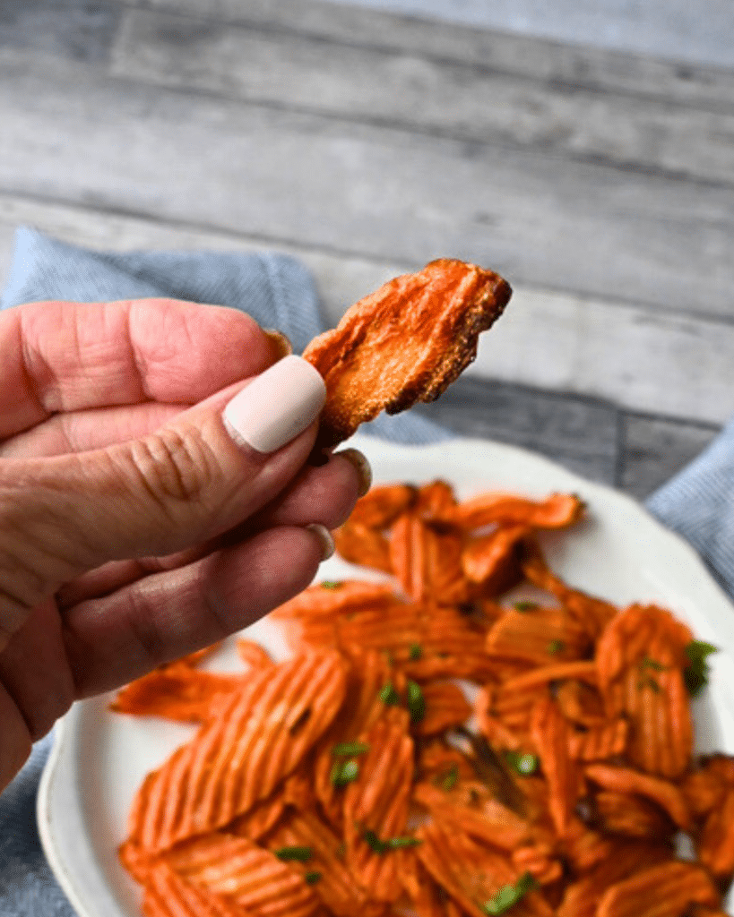 A hand with pale nail polish lifts a crinkle-cut carrot chip over a herb-garnished bowl of chips on a wooden table.