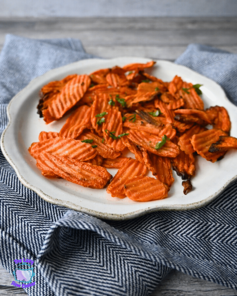 Crinkle-cut carrot chips topped with herbs are served on a white plate over a blue and white cloth.