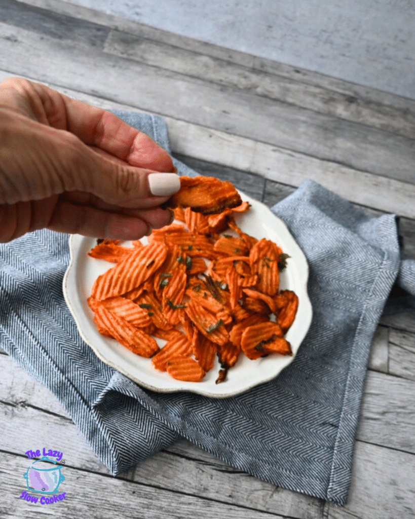 A hand with pale nail polish lifts a crinkle-cut carrot chip over a herb-garnished bowl of chips on a wooden table.