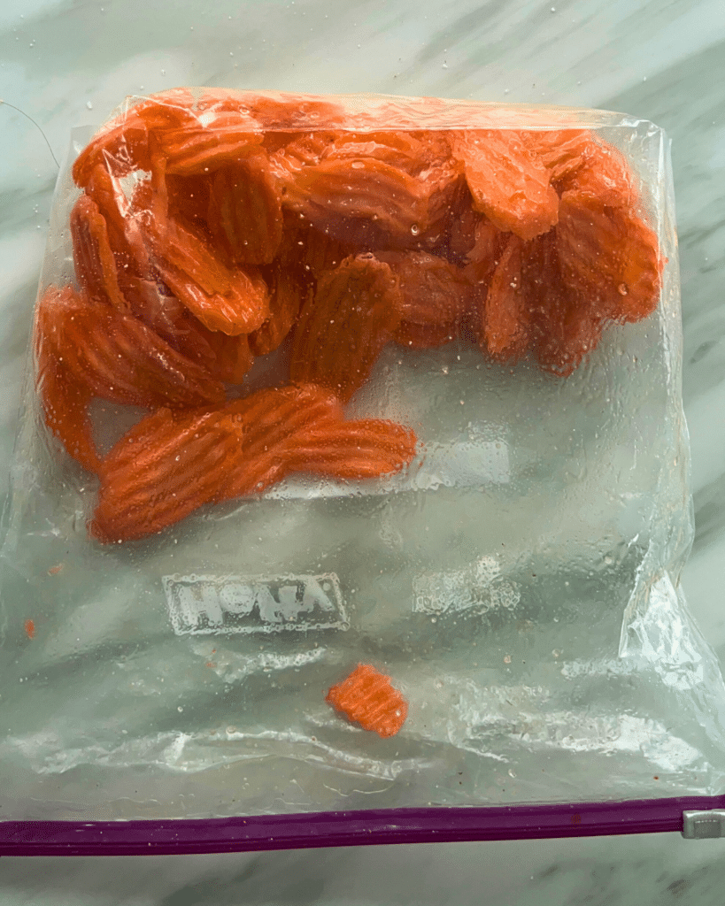 A clear ziplock bag filled with crinkle-cut carrot slices rests on a pale marble countertop, ready for cooking or meal prep.