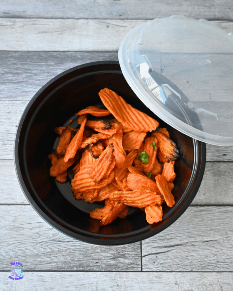 Orange crinkle-cut chips rest in a black bowl with a clear lid, placed on a wooden table.