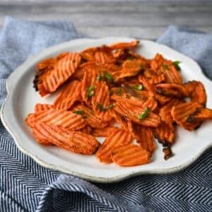 Crinkle-cut carrot chips topped with herbs are served on a white plate over a blue and white cloth. Text reads: Air Fryer Carrot Chips.