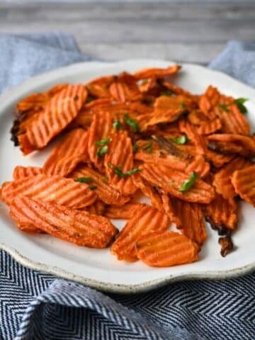 Crinkle-cut carrot chips topped with herbs are served on a white plate over a blue and white cloth. Text reads: Air Fryer Carrot Chips.