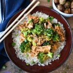 A bowl of white rice with shredded slow cooker chicken and broccoli, and sesame seeds, served with chopsticks. Roasted potatoes and a blue napkin are beside it.
