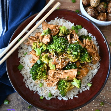 A bowl of white rice with shredded slow cooker chicken and broccoli, and sesame seeds, served with chopsticks. Roasted potatoes and a blue napkin are beside it.