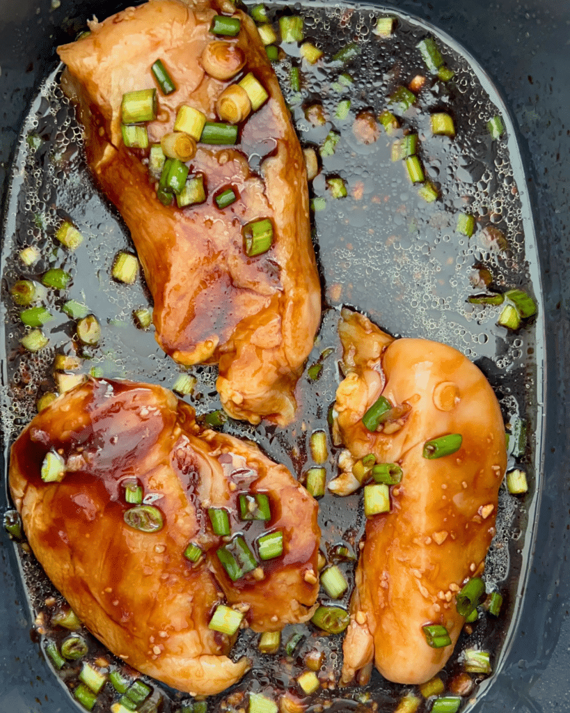 Three raw chicken breasts and sauce in a slow cooker