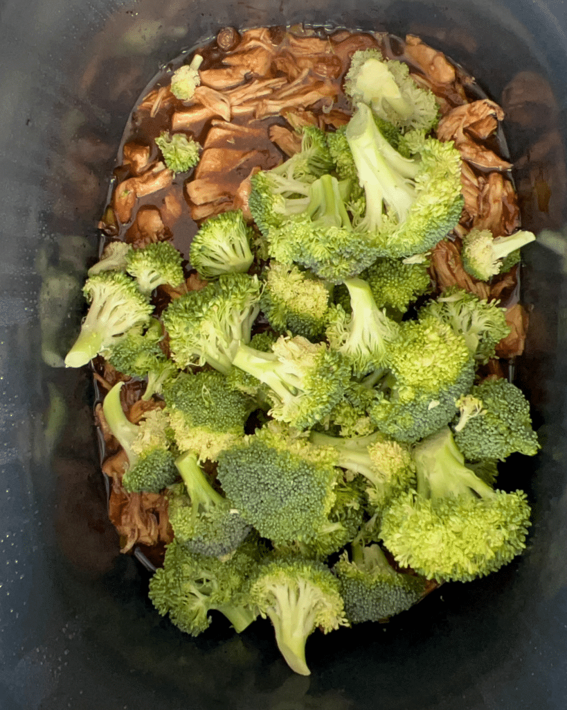 Broccoli florets sit atop shredded meat covered in brown sauce inside a slow cooker, about to be mixed and cooked together.