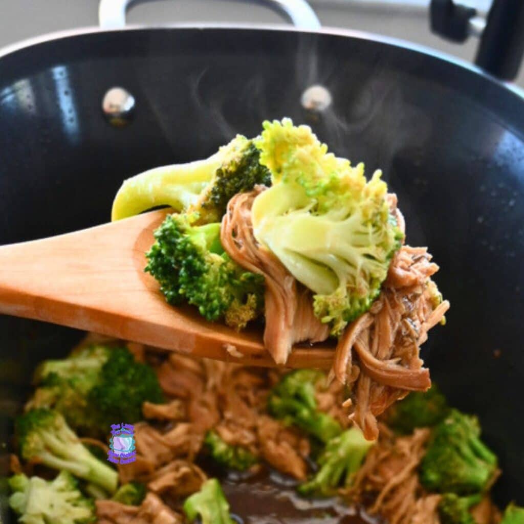 A wooden spoon lifts shredded chicken and broccoli from a steaming black slow cooker with savory sauce, showing a chicken and broccoli recipe