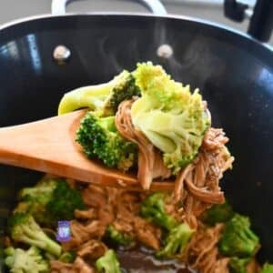 A wooden spoon lifts shredded chicken and broccoli from a steaming black slow cooker with savory sauce, showing a chicken and broccoli recipe