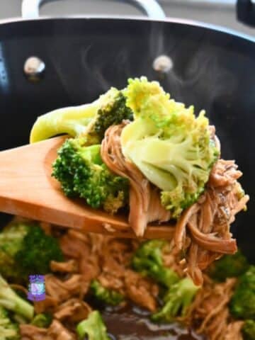 A wooden spoon lifts shredded chicken and broccoli from a steaming black slow cooker with savory sauce, showing a chicken and broccoli recipe