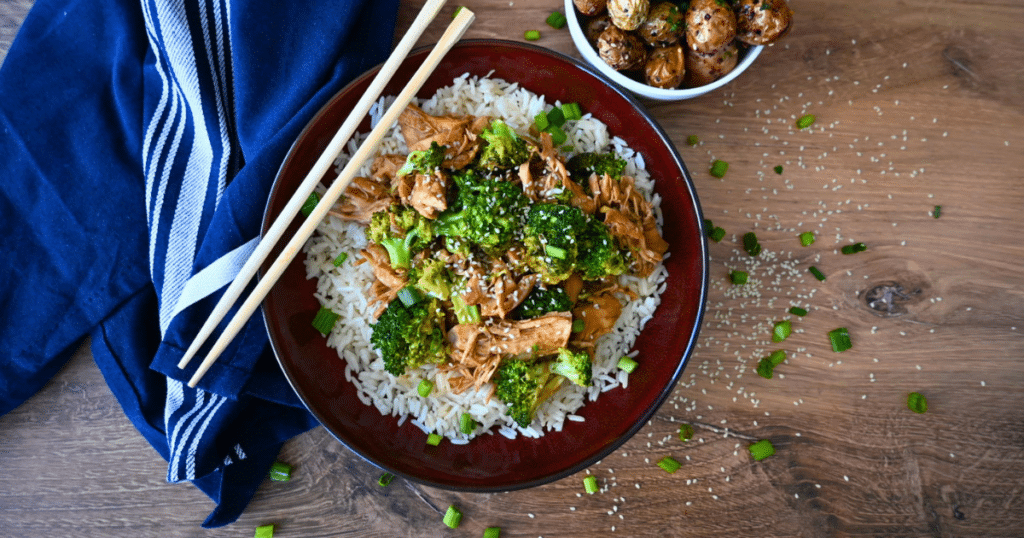 A bowl of white rice with shredded slow cooker chicken and broccoli, and sesame seeds, served with chopsticks. Roasted potatoes and a blue napkin are beside it.