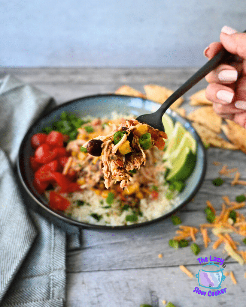 A hand holds a forkful of slow cooker shredded chicken Santa Fe with corn, and beans above a rice bowl with tomatoes, lime, cheese, and chips.