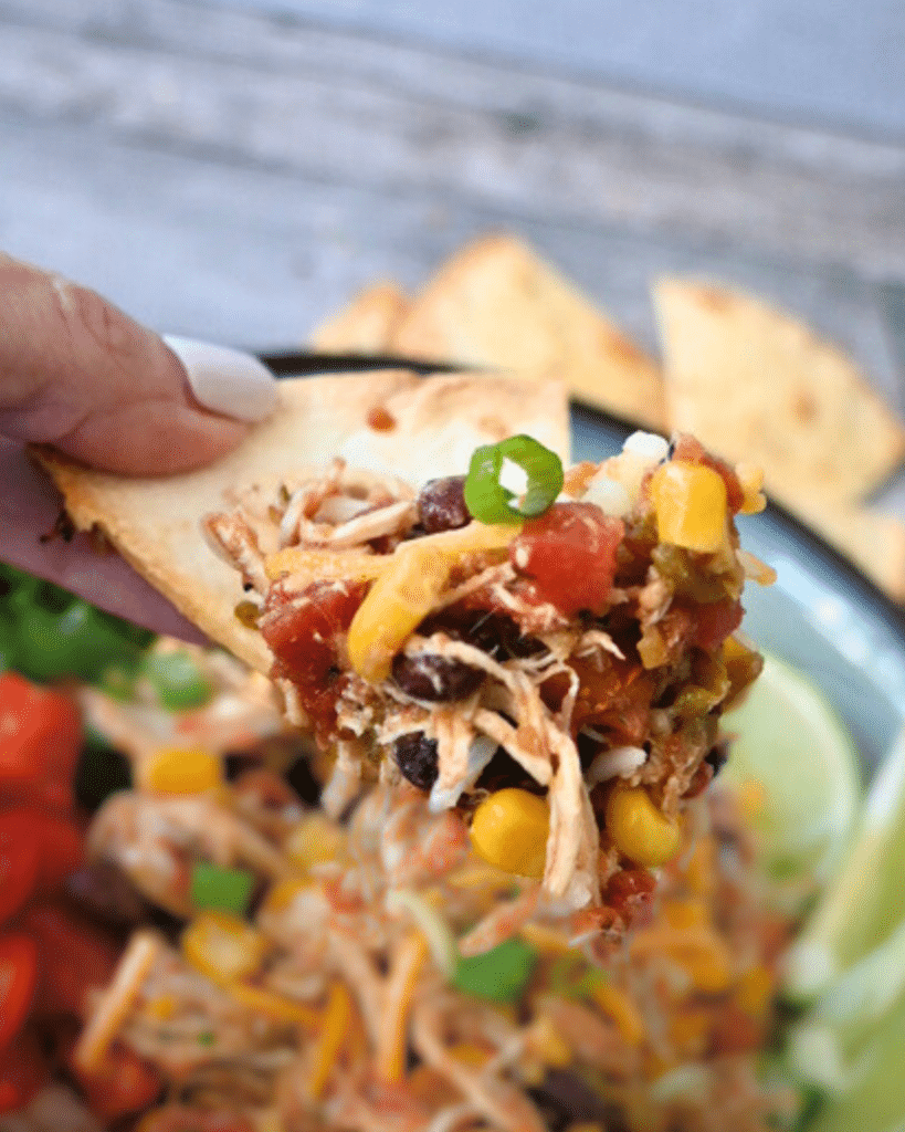 A hand holds a chip with shredded slow cooker Santa Fe chicken, corn, black beans, and green onions; more is in a bowl nearby.