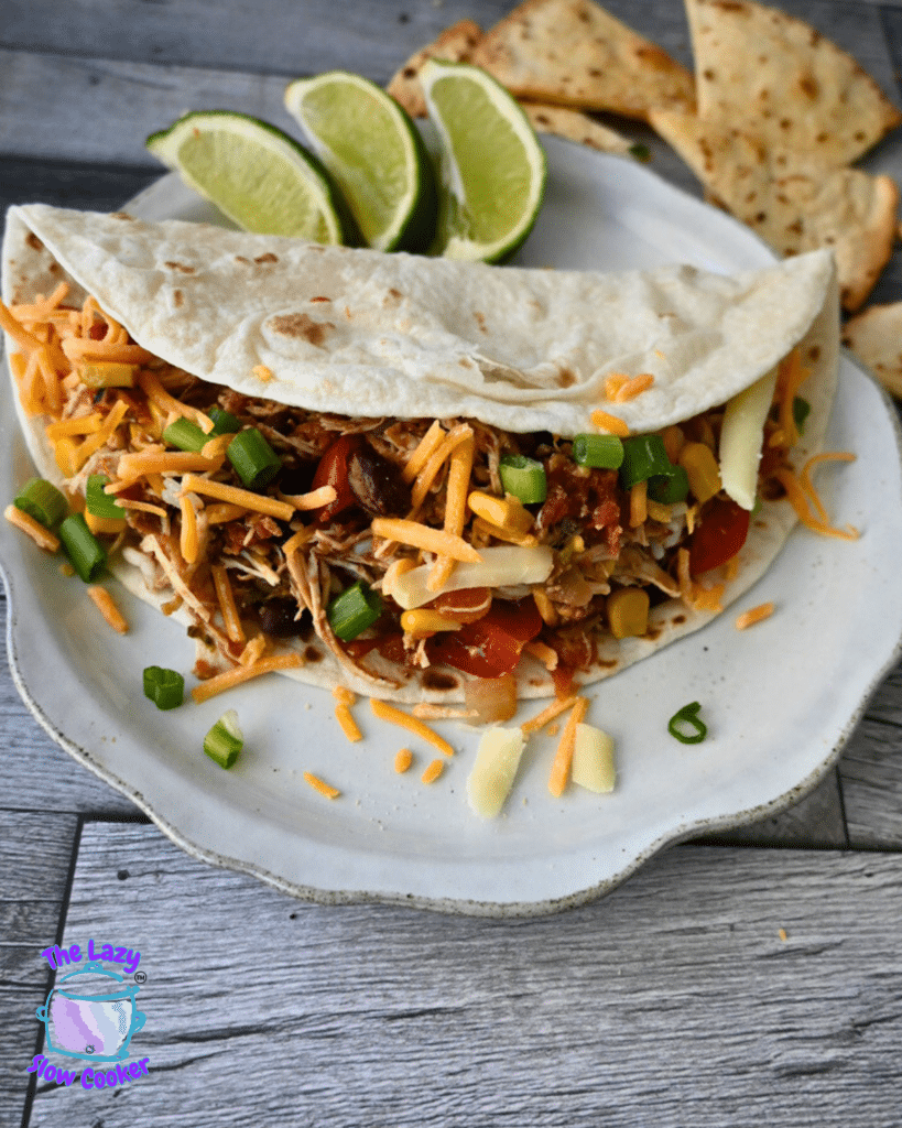 A plate with a shredded Santa Fe chicken taco, topped with cheese, tomatoes, and green onions, served with lime wedges and tortilla chips.