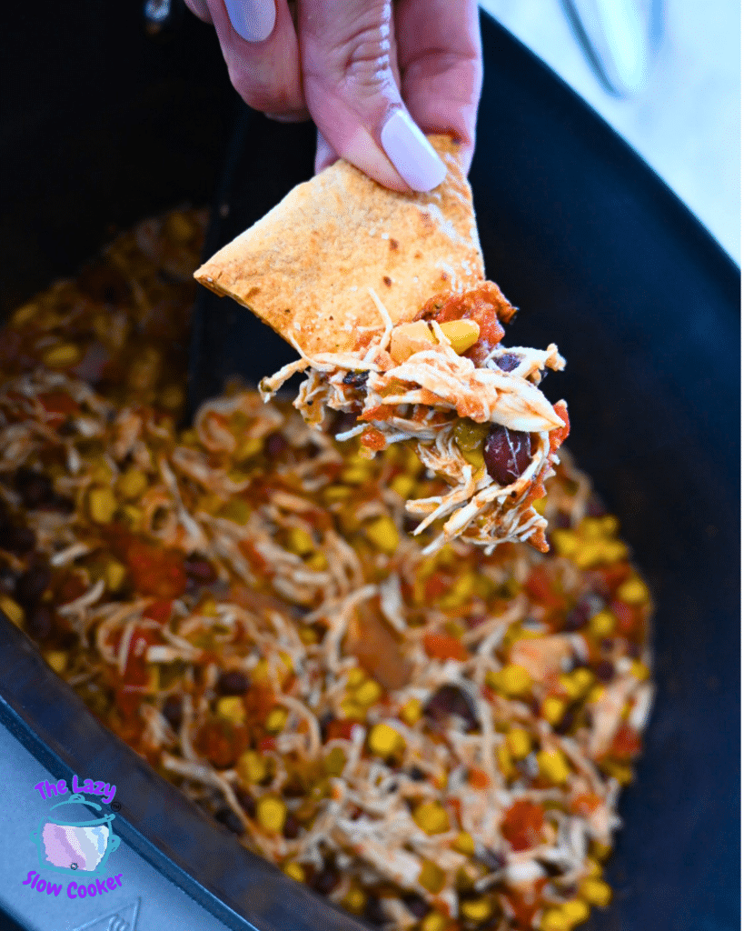 A hand holds a chip with shredded slow cooker Santa Fe chicken, corn, and black beans over a slow cooker filled with the same