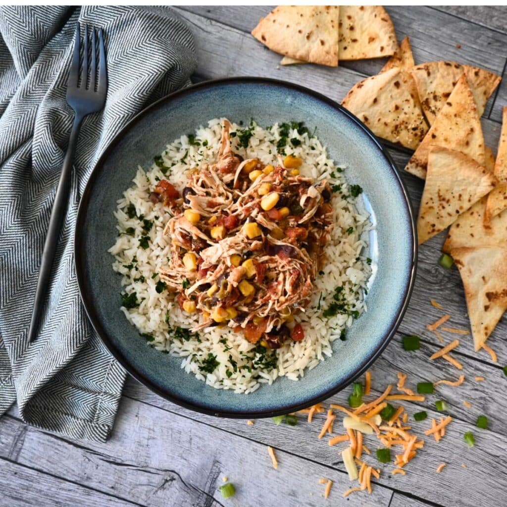 A bowl of slow cooker shredded chicken with corn, black beans, and rice, topped with cheese and green onions, served with tortilla chips.