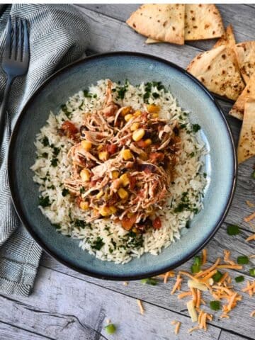 A bowl of slow cooker shredded chicken with corn, black beans, and rice, topped with cheese and green onions, served with tortilla chips.