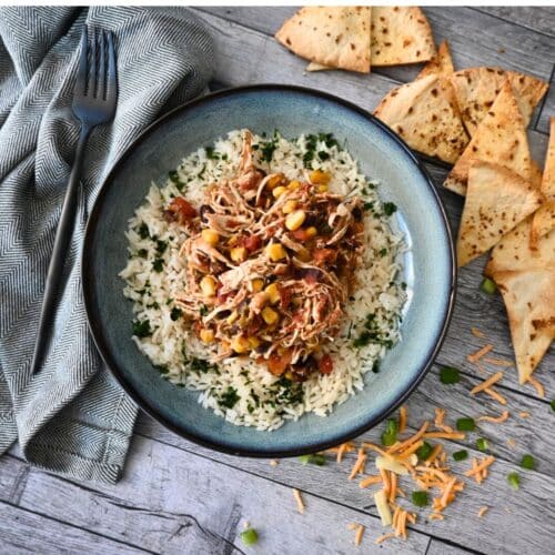 A bowl of slow cooker shredded chicken with corn, black beans, and rice, topped with cheese and green onions, served with tortilla chips.