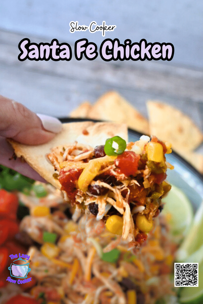 A hand holds a chip with shredded slow cooker Santa Fe chicken, corn, black beans, and green onions; more is in a bowl nearby.