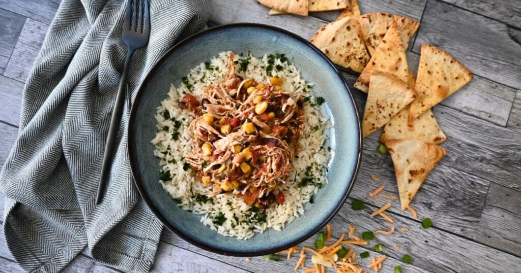 A bowl of slow cooker shredded chicken with corn, black beans, and rice, topped with cheese and green onions, served with tortilla chips.