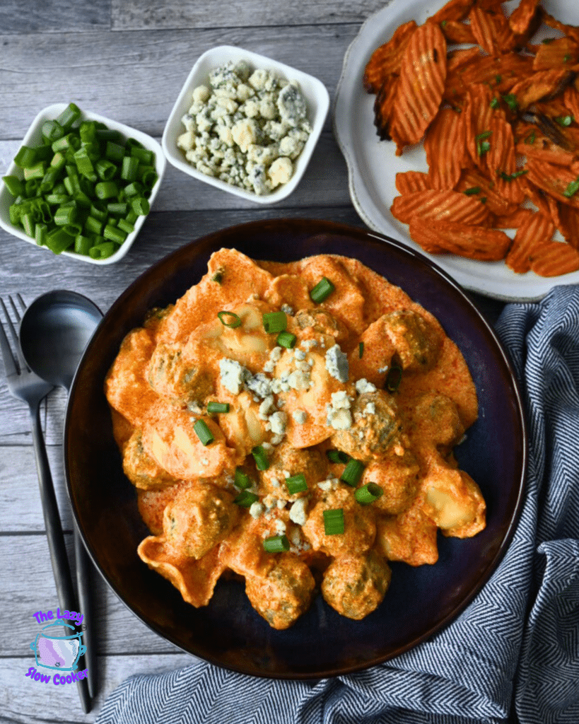 A bowl of creamy slow cooker pasta with meatballs, topped with blue cheese and green onions, served with crinkle-cut air fried carrot chips on the side