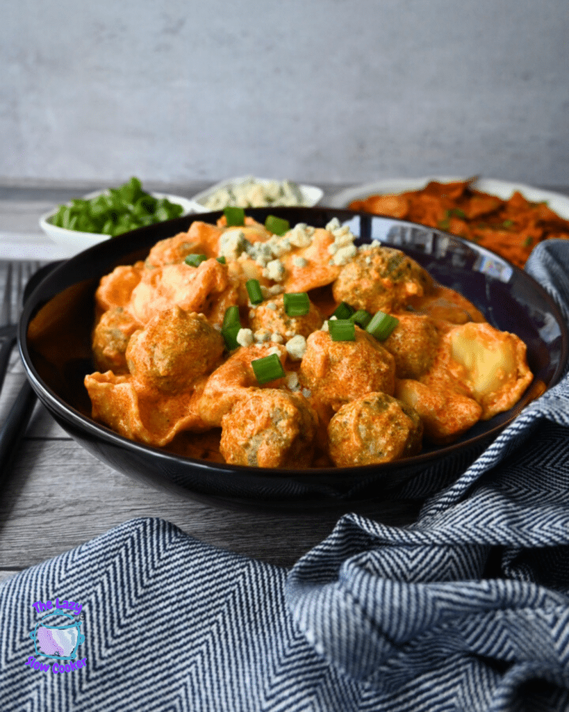 A black bowl of creamy pasta and meatballs, topped with green onions and cheese, sits on a blue-striped cloth with extra toppings nearby.