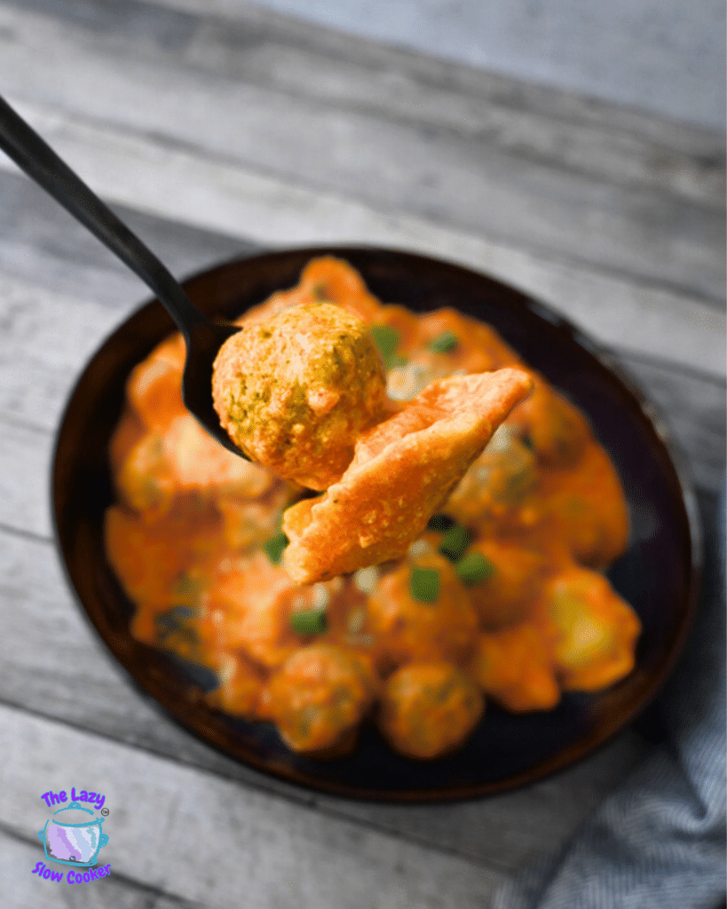 A fork with a meatball and tortellini hovers over a bowl of slow cooker meatballs in creamy orange sauce, topped with fresh herbs.