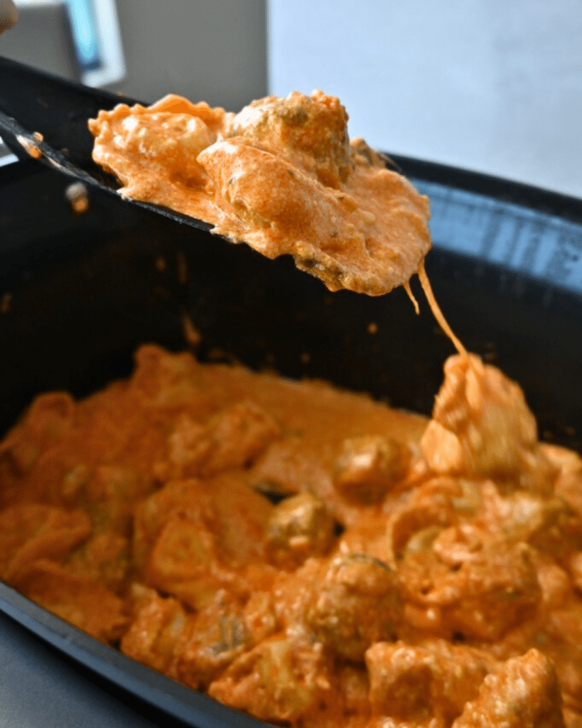 A spoon lifts saucy, buffalo sauce coated meatballs from a slow cooker filled with more meatballs and tortellini in thick, creamy sauce.
