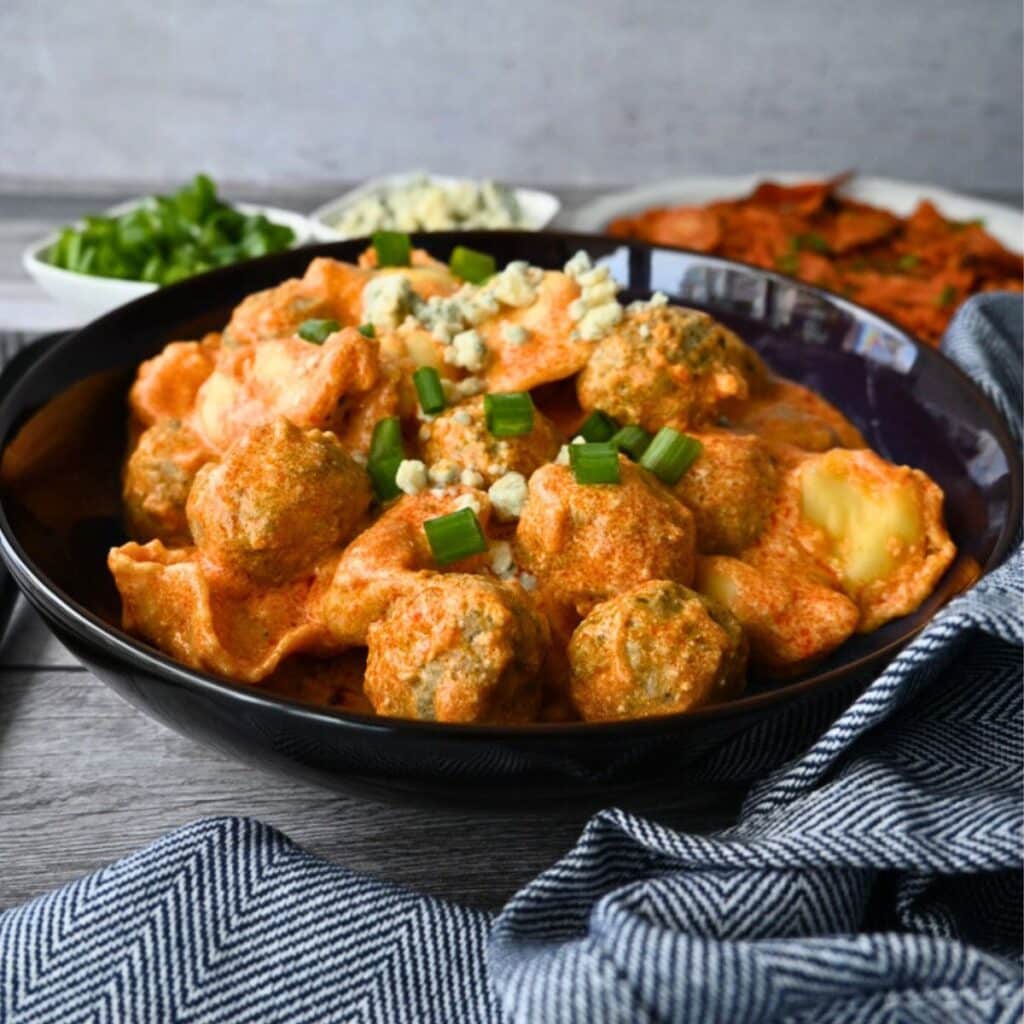 A black bowl of creamy pasta and meatballs, topped with green onions and cheese, sits on a blue-striped cloth with extra toppings nearby.