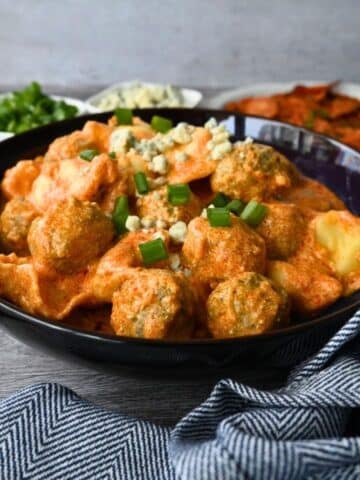 A black bowl of creamy pasta and meatballs, topped with green onions and cheese, sits on a blue-striped cloth with extra toppings nearby.