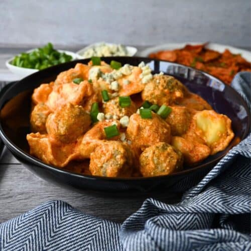A black bowl of creamy pasta and meatballs, topped with green onions and cheese, sits on a blue-striped cloth with extra toppings nearby.