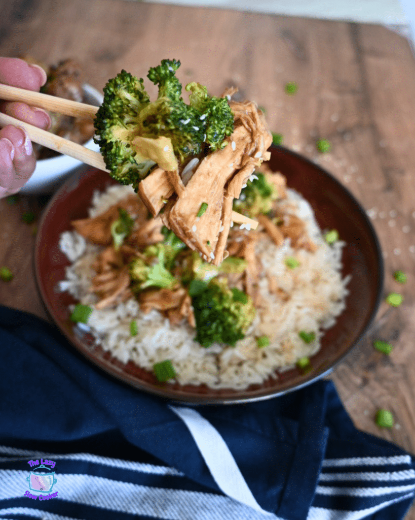 Chopsticks lift broccoli and shredded chicken over a plate of rice with slow cooker chicken, broccoli, sauce, and green onions.