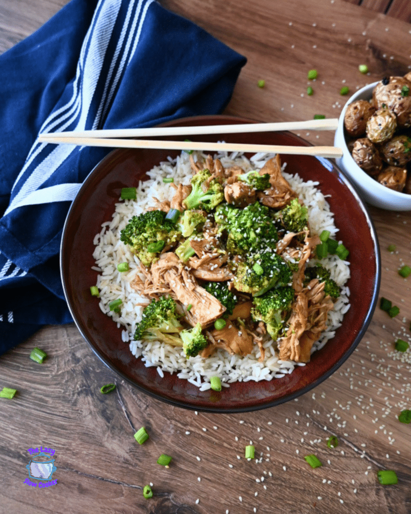 A bowl of white rice with shredded slow cooker chicken and broccoli, and sesame seeds, served with chopsticks. Roasted potatoes and a blue napkin are beside it.