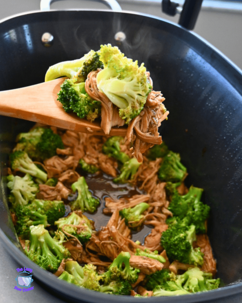 A wooden spoon lifts shredded chicken and broccoli from a steaming black slow cooker with savory sauce, showing a chicken and broccoli recipe