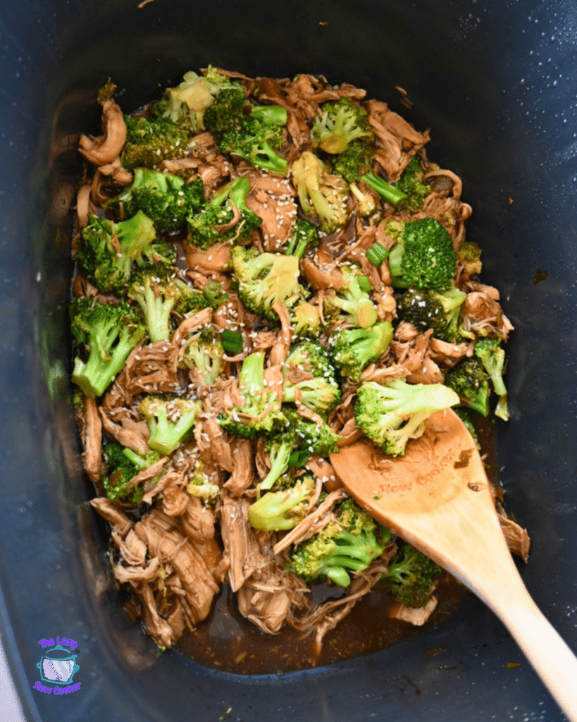 Slow cooker shredded beef and broccoli in sauce, sprinkled with sesame seeds. A wooden spoon rests in the mixture, ready to serve.