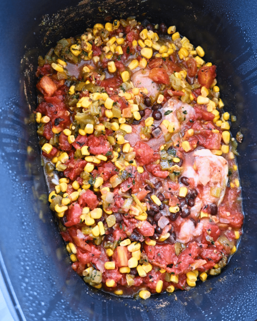 A slow cooker with uncooked chicken, corn, black beans, diced tomatoes, and vegetables mixed together in a savory sauce.