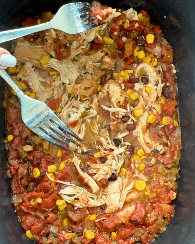 Two forks pull apart cooked chicken in a slow cooker filled with diced tomatoes, corn, black beans, and spices for a hearty dish.