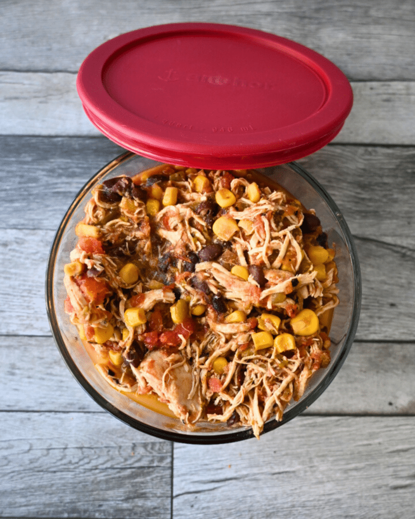 A glass bowl with a red lid holds leftover santa fe slow cooker shredded chicken with corn, black beans, and diced tomatoes on a wooden table.