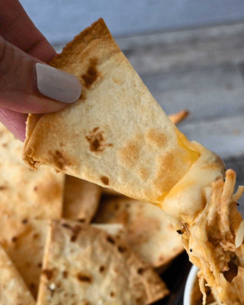 A person with light-painted nails holds a cheesy tortilla chip, with more chips behind it, ready to enjoy as nachos.