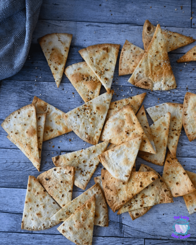 Golden-brown homemade air fryer pita chips, cut into triangles and seasoned, are spread out on a rustic wooden table.