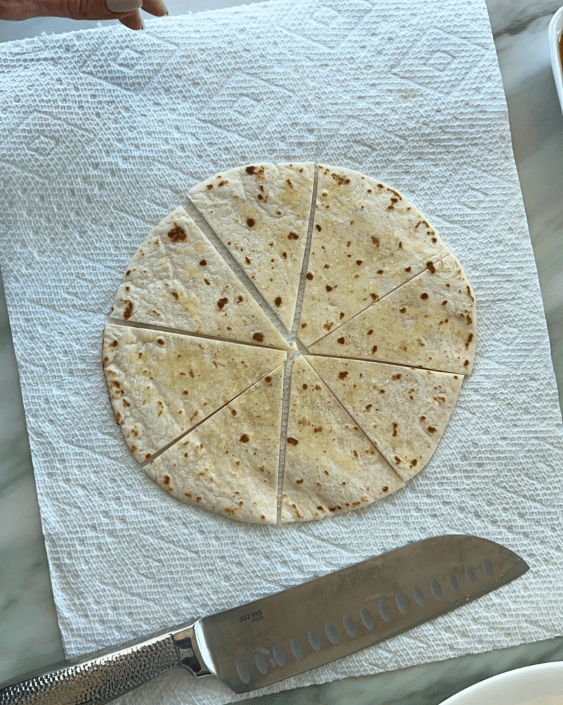 A tortilla, sliced into eight wedges for a recipe, rests on a paper towel beside a knife. A hand appears at the top edge.