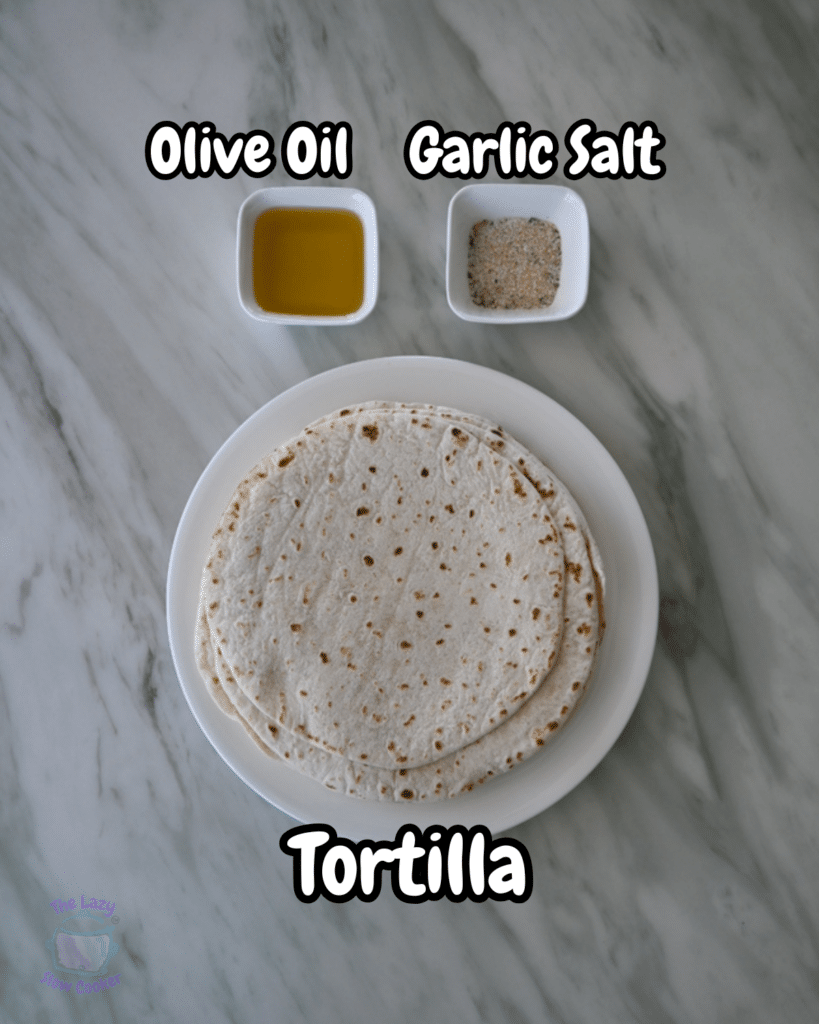 Stacked tortillas sit on a plate atop a marble counter, with labeled dishes of olive oil and garlic salt arranged above them.