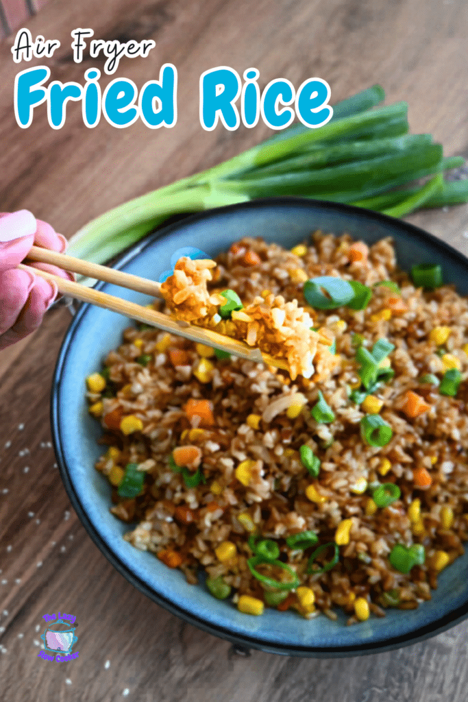A hand lifts a bite of air fryer fried rice with chopsticks from a bowl on a wooden table; fresh green onions are nearby.