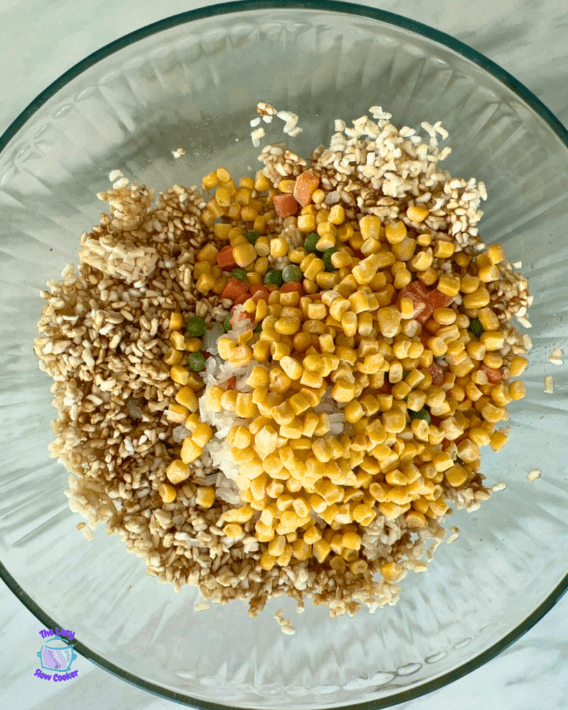 A glass bowl holds uncooked rice, frozen corn, peas, carrots, and seasonings, ready to mix for air fryer fried rice.