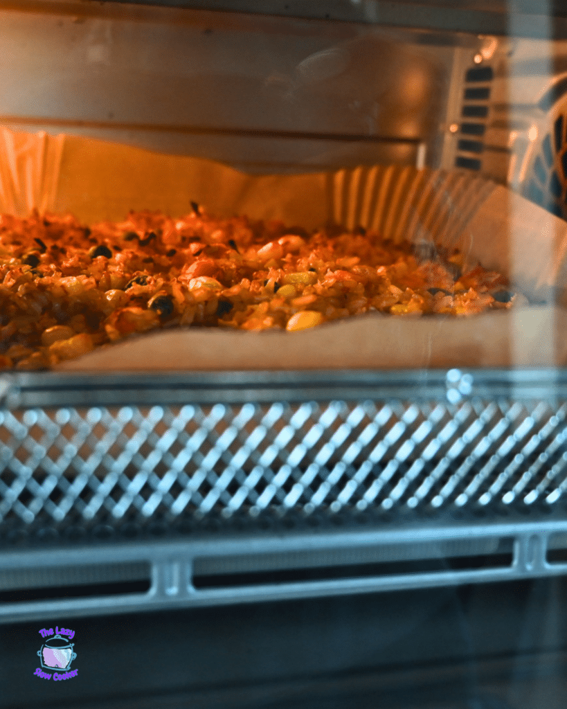 Through an oven door, a tray of air fryer fried rice with vegetables bakes on parchment, edges turning golden and crisp.