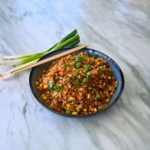 A bowl of air fryer fried rice with mixed vegetables and green onion garnish sits on marble, next to chopsticks and fresh green onions.