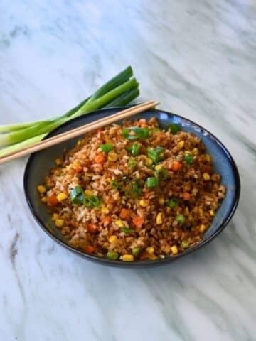 A bowl of air fryer fried rice with mixed vegetables and green onion garnish sits on marble, next to chopsticks and fresh green onions.